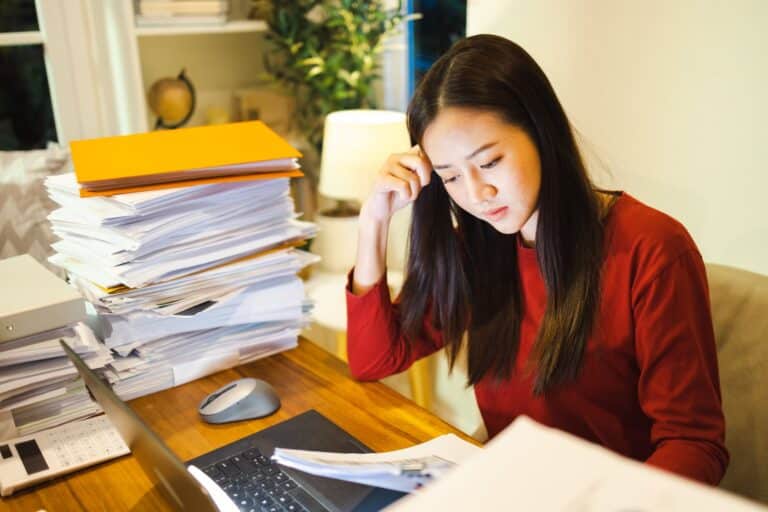 Woman at a desk with a large stack of papers, looking focused and overwhelmed