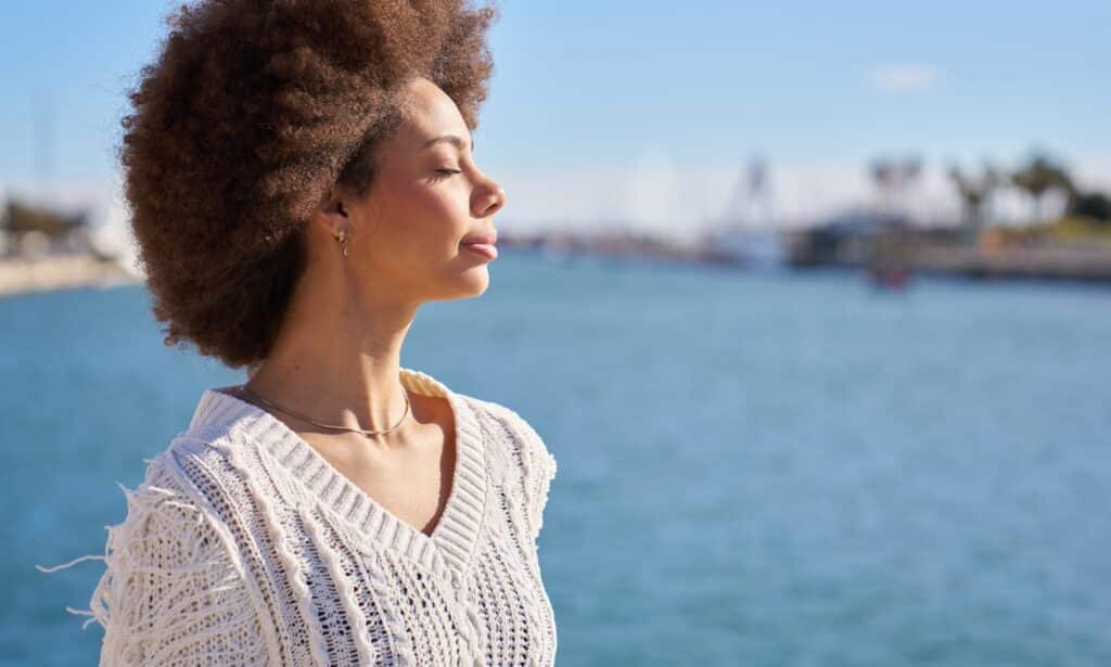 Woman with curly hair standing by the water with eyes closed, looking calm and centered