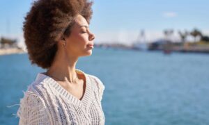 Woman with curly hair standing by the water with eyes closed, looking calm and centered