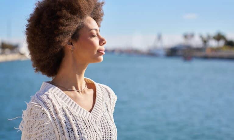 Woman with curly hair standing by the water with eyes closed, looking calm and centered
