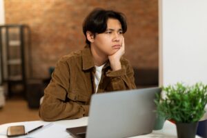 Pensive young adult resting their chin on their hand at a desk with a laptop