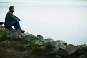 Man sitting on rocks by the water, looking out thoughtfully